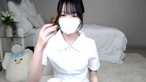 Media: Video of a young Asian woman with straight black hair, wearing a white dress, face mask, and holding a hair tie, seated on a plush bear in a minimalist, white bedroom.