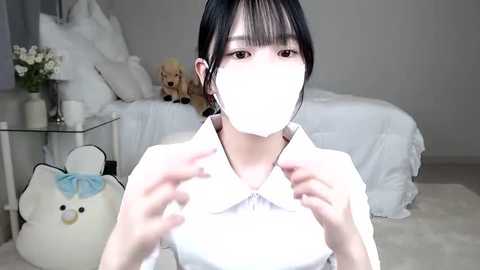 Media: Video of an Asian woman in a white mask, white blouse, and light blue bow, sitting on a beige rug in a minimalist bedroom with a white bed, teddy bear, and vase.