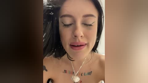 Media: Video of a close-up of a young woman with black hair, wearing black eyeliner and a silver necklace. She has a tattoo on her collarbone and a heart-shaped piercing on her lip.