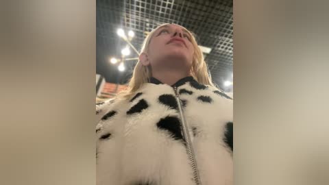 Media: Video of a young Caucasian woman with straight blonde hair, wearing a black-and-white cow print jacket, looking upwards in a brightly lit indoor setting with a grid ceiling and blurred background.