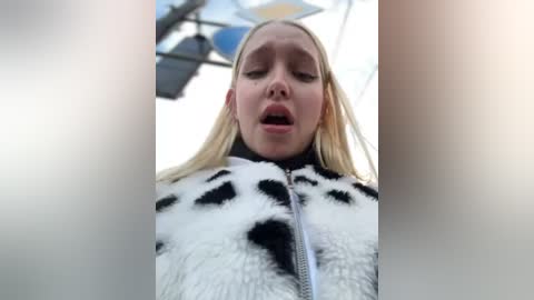 Media: A video of a young blonde woman with closed eyes and open mouth, wearing a white, fuzzy jacket with black panda-like patterns, against a blurry outdoor backdrop.