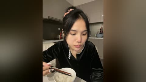 Media: Video of an Asian woman with long black hair in a ponytail, wearing a black velvet jacket, eating noodles with chopsticks in a modern kitchen with light-colored cabinets and a tiled backsplash.