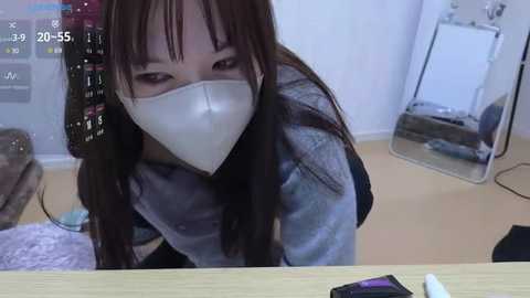 Media: Video of a young Asian woman with long brown hair, wearing a white face mask, sitting on a bed, looking directly at the camera. Background shows a mirror and a digital clock.