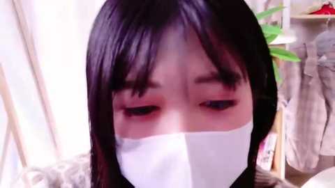 Media: Video of an Asian woman with long black hair, wearing a white surgical mask, looking down with a sad expression. Background includes a green plant and hanging clothes in a room.