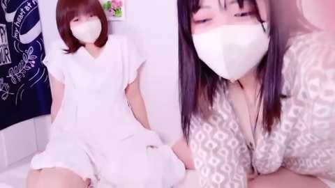 Media: A video features two Asian women with fair skin, one in a white dress, the other in a patterned blouse. Both wear face masks, with one smiling and the other looking serious.