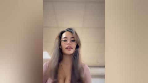 Media: Video of a young woman with long, brown hair, wearing glasses and a light pink top, standing in a beige room with a tiled ceiling.