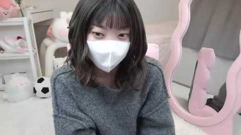 Media: Video of an Asian woman with shoulder-length dark hair, wearing a gray sweater, white mask, and pink chair in a pastel-colored, cluttered room with toys and a soccer ball.