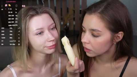 Media: Video of two young women, one blonde and one brunette, eating a banana in a modern, dimly-lit room with a television showing a fitness app interface in the background.