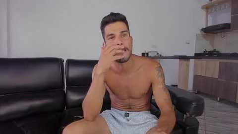 Media: Video of a shirtless, muscular, tan-skinned man with short dark hair and a beard, wearing light blue shorts, sitting on a black leather sofa in a modern kitchen with wooden cabinets.