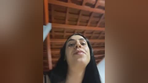 Media: A video of a young woman with long black hair, wearing minimal makeup, standing under a wooden ceiling with visible beams, indoors.