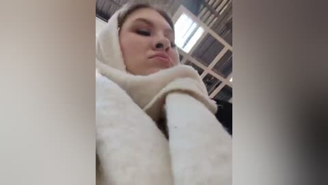 Media: A video of a young woman with fair skin, wearing a white hooded sweater and a pink face mask, taken indoors with a corrugated metal ceiling and bright light streaming through windows.