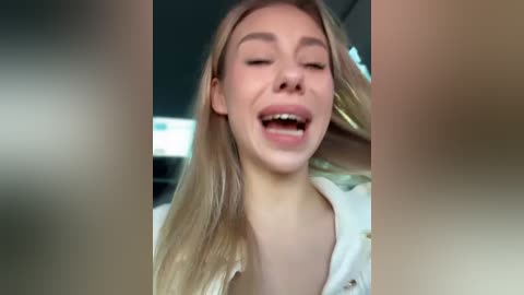 Media: A close-up video of a smiling, light-skinned young woman with long blonde hair, wearing a white shirt, captured in a blurry car interior.