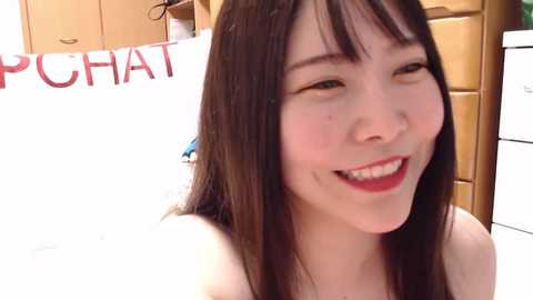 Media: Video of a smiling East Asian woman with straight black hair and fair skin, wearing red lipstick, in a room with wooden cabinets and a \"CHAT\" sign.