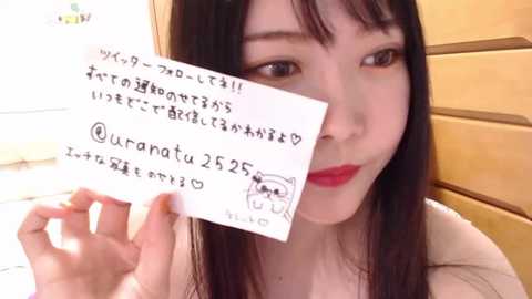 Media: Video of an Asian woman with long black hair, fair skin, and red lipstick, holding a handwritten note with Japanese text and a drawing, smiling.