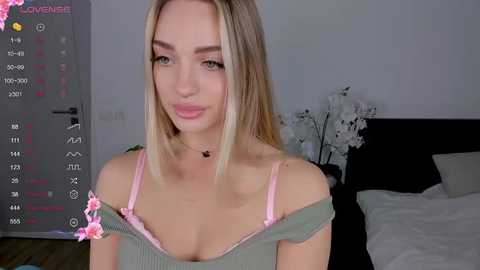 Media: Video of a young Caucasian woman with straight blonde hair, light skin, and a slender physique, wearing a pink bra and green off-shoulder top, in a bedroom with a calendar, white flowers, and a bed.