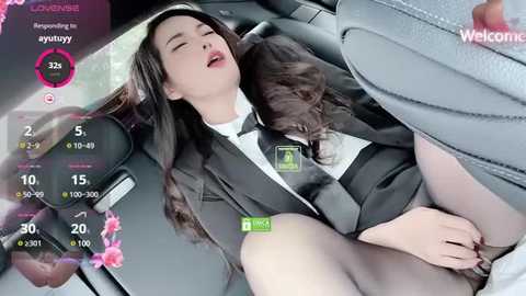 Media: Video of a young Asian woman with long hair, wearing a black suit, lying in a car, partially undressed, with a man's hand on her thigh, surrounded by digital game interface elements.