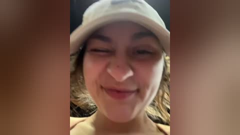 Media: Video of a young woman with fair skin and curly brown hair, wearing a beige hat, smiling, with blurred background.