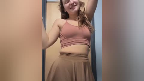 Media: Video of a young, light-skinned woman with curly brown hair, wearing a pink crop top and beige skirt, standing in a doorway, smiling, arms raised.