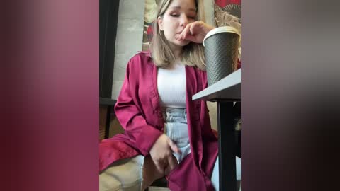 Media: Video of a young Asian woman with straight, shoulder-length blonde hair, wearing a pink robe over a white top and high-waisted blue jeans, sitting in a cafe, holding a coffee cup.