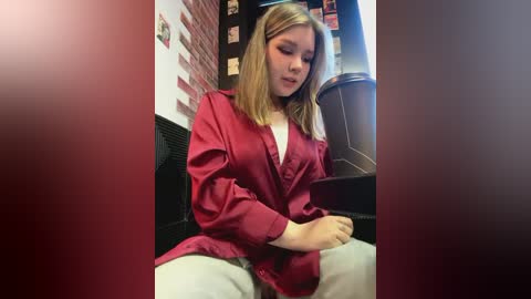 Media: Video of a young woman with shoulder-length blonde hair, wearing a red jacket, seated in a dimly lit room with brick walls and a bulletin board.