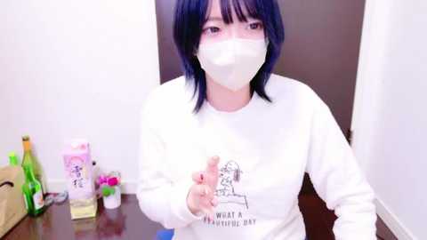 Media: Video of a woman with short dark hair and a white mask, wearing a white sweater with \"What a Wonderful World\" text, pointing at a wall. Background includes a desk with various personal items.