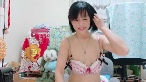 Media: Video of an East Asian woman with short black hair, fair skin, and small breasts, wearing a pink lace bra, smiling while adjusting her hair. Background features a colorful, cluttered room with stuffed toys and a wicker basket.