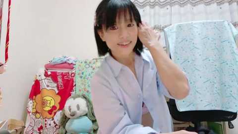 Media: A video of a young woman with shoulder-length black hair, smiling, wearing a light blue shirt, sitting in a cluttered bedroom with colorful pillows, stuffed animals, and a blue towel.