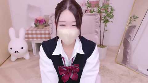 Media: Video of an Asian girl in a white shirt, black vest, and red tie, wearing a beige mask, kneeling in a soft, beige room with bunny decor, pink flowers, and a white chair.