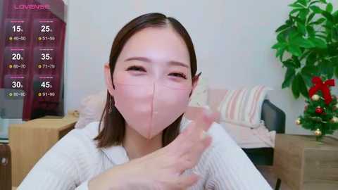 Media: A video of an Asian woman with straight, shoulder-length brown hair, wearing a white mask and a white knitted sweater, sitting in a living room with a potted plant and a striped cushion on a couch.