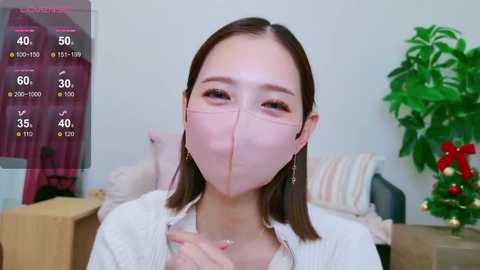 Media: Video of an East Asian woman in a white mask, wearing a white blouse, sitting in a room with a plant, pink curtains, and a wooden dresser.