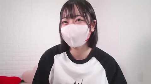 Media: Video of an East Asian woman with shoulder-length black hair, wearing a white face mask, black and white top, and a red object in the background against a white brick wall.