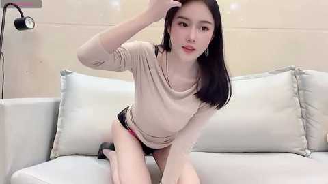 Media: A video of an East Asian woman with straight black hair, fair skin, and a petite frame, wearing a beige top and black shorts, kneeling on a light-colored sofa. The background shows a minimalist interior with beige walls and a black lamp.