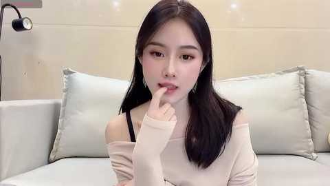 Media: Video of an East Asian woman with long black hair, fair skin, and brown eyes, wearing a beige off-shoulder sweater, seated on a light gray couch, finger to her lips, in a minimalist, modern living room.