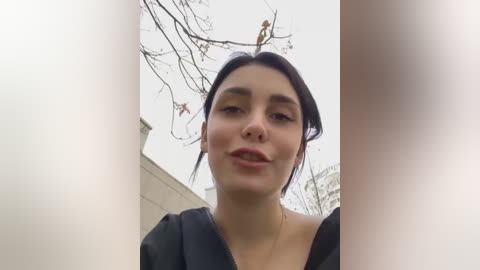 Media: A video of a young woman with fair skin and dark hair, wearing a black jacket, standing outside with bare tree branches and a cloudy sky in the background.