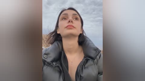 Media: A video of a young woman with medium skin tone, brown hair, and full lips, wearing a black puffy jacket, looking upward against a cloudy sky.