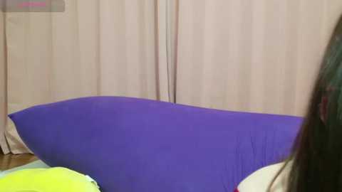 Media: A video of a child's bedroom with a purple beanbag chair against a beige curtain backdrop, showing a cluttered space with a green and yellow pillow visible on the left.