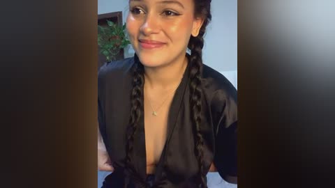 Media: Video of a smiling Latina woman with medium skin tone, wearing black braided hair, a black satin robe, and a delicate necklace. Background includes a green plant and light-colored wall.