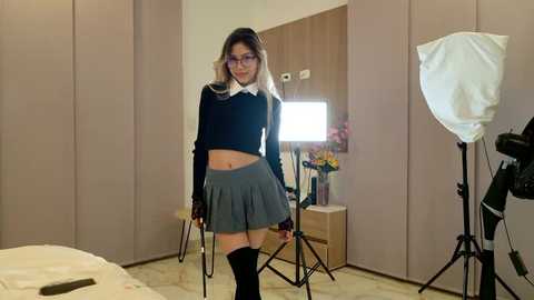 Media: Video of a petite, East Asian woman with long, straight, black hair, wearing a black sweater, grey pleated skirt, and knee-high black socks, standing in a modern, minimalist bedroom with beige walls, wooden closet, and a large studio light.