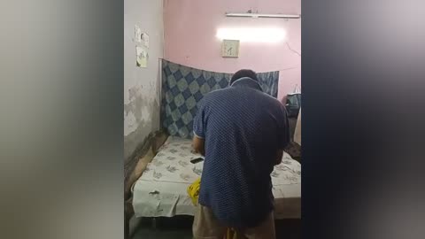 Media: Video of a small, dimly lit room with peeling pink walls and a blue-patterned bedspread. A man with short black hair and a blue shirt is seen from behind, bending over.