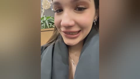 Media: Video of a young woman with fair skin and brown hair, smiling warmly, wearing a light gray blazer over a beige top, in an office setting with a potted plant and wooden furniture in the background.