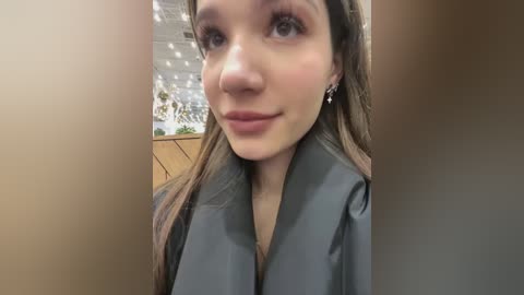 Media: Video of a young Caucasian woman with fair skin and long brown hair, wearing a black robe. She has a serene expression with subtle makeup and is surrounded by blurred, warm-toned background.