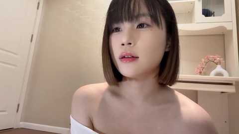 Media: Video of a young Asian woman with short, straight black hair, light skin, and a petite frame, wearing a white off-shoulder top, sitting indoors near a closed door and beige shelves.