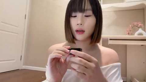 Media: Video of a young Asian woman with straight, shoulder-length black hair and pale skin, wearing a white off-shoulder top. She is applying eyeliner in a bathroom with beige walls, a wooden vanity, and a closed white door.