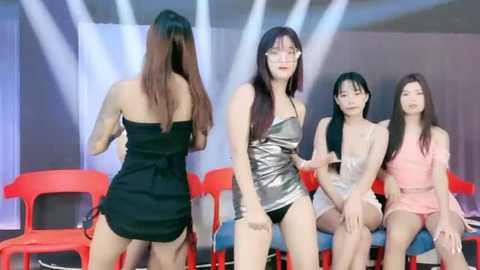 Media: Video of three Asian women in revealing outfits, seated on red chairs; one in a black dress, one in a metallic dress, and one in a pink dress.