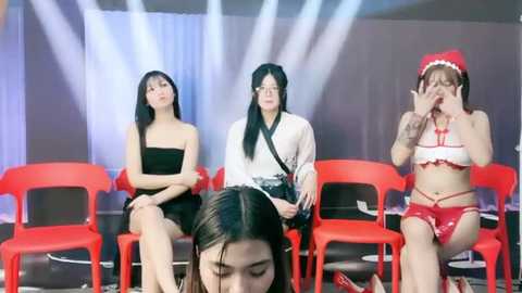 Media: Video of four women in a studio, wearing diverse outfits: black dress, white kimono, red lingerie, and Santa hat. They sit on bright red plastic chairs in front of a dimly lit background with spotlights.