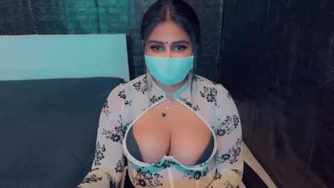 Media: A video of a woman with dark skin, large breasts, and a face mask, wearing a white floral-patterned blouse with a plunging neckline, in a dimly lit room.