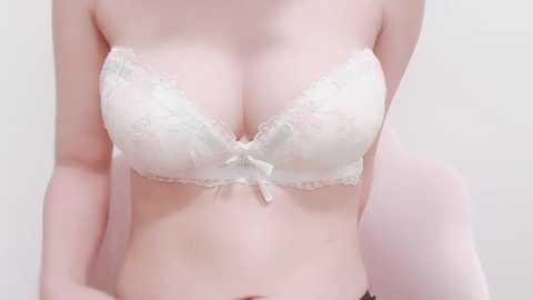 Media: Video of a light-skinned woman wearing a white lace bra with a small bow center. Her breasts are moderately large, and she is standing against a plain, light-colored background.