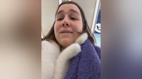 Media: Video of a young woman with light skin and straight brown hair, wearing a fluffy purple and white bathrobe, smiling slightly. The background shows a white wall with a blurred blue sign.