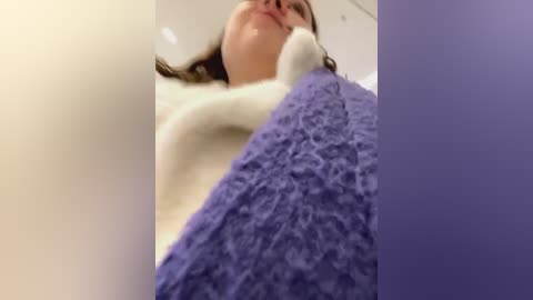 Media: A video captures a woman in a white sweater, with a purple knit blanket covering her lower body, lying on a light-colored couch. Her face is partially visible, smiling with glasses.