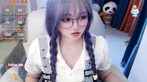 Media: Video of a young Asian woman with purple hair in braids, wearing glasses, a white blouse, and overalls, sitting in a chair, surrounded by plush toys.